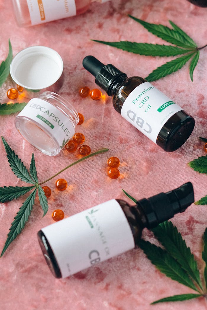 A variety of CBD oils and capsules surrounded by cannabis leaves on a pink background.